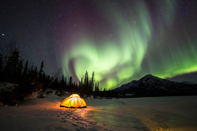 Green aurora borealis over an illuminated yellow tent by a frozen river at night.