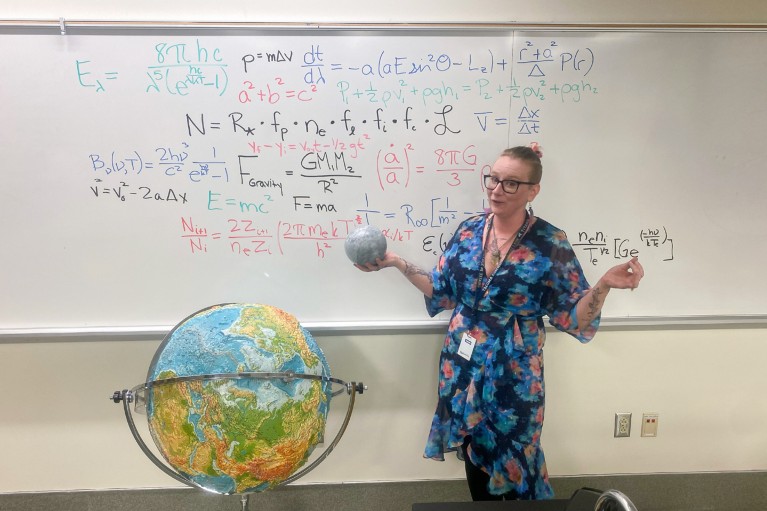 Professor Seven Rasmussen teaches in front of a whiteboard covered with equations, while standing next to a model earth and holding a model moon