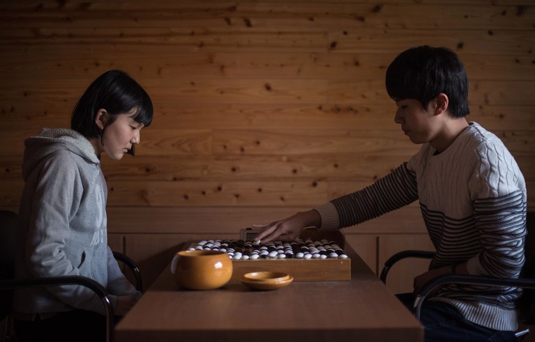 Two people sit opposite each other at a wooden table playing Go.