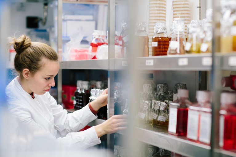 A young woman in a lab coat looking at bottles on chemicals on shelves in a store room