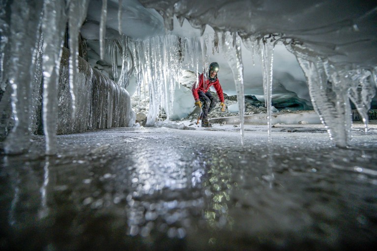 A man wearing a helmet with a head torch crouches while moving through an icy cave with many icicles