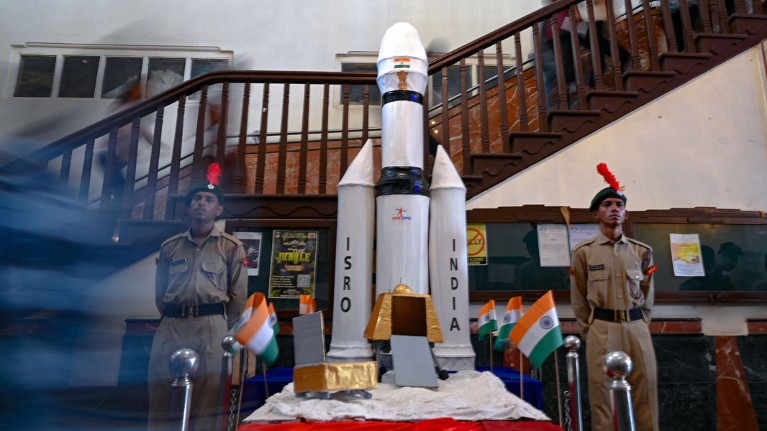 Students in a Mumbai college walk past a model of India's Chandrayaan-3 spacecraft and its two uniformed guards.