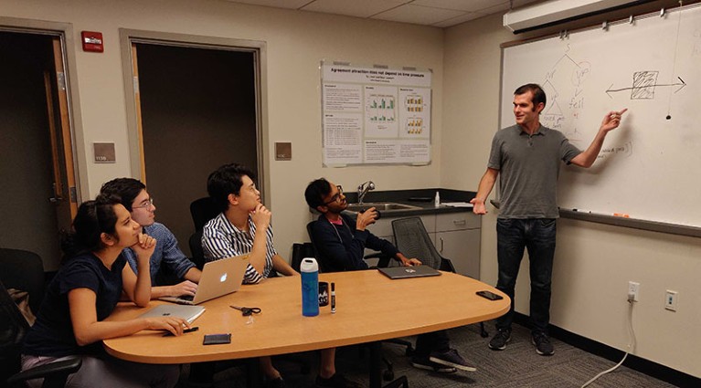 Person standing in front of whiteboard looking towards four people sitting at a table