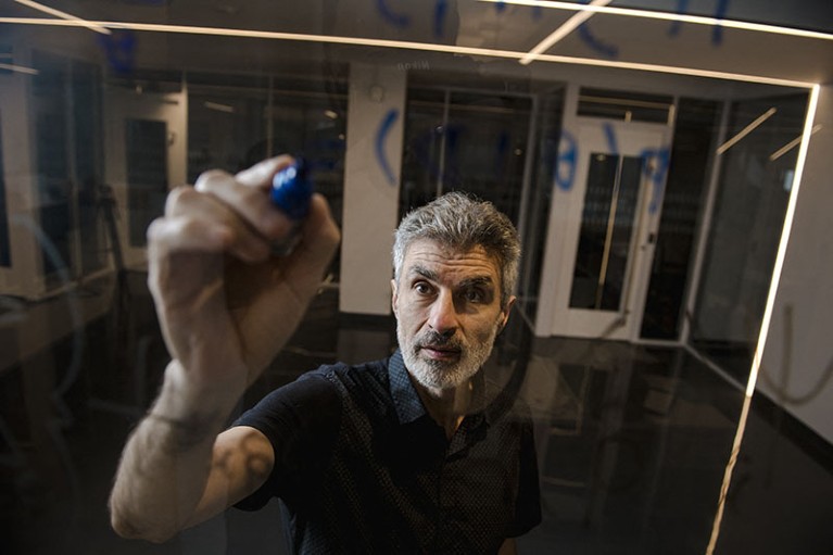 Person with grey hair and beard facing towards camera, holding pen writing on glass