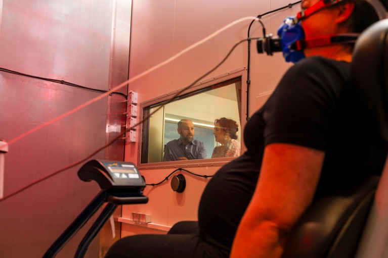 Researchers observe a pregnant woman in climate chamber flooded with red light wearing a mask and cycling on a cycling machine