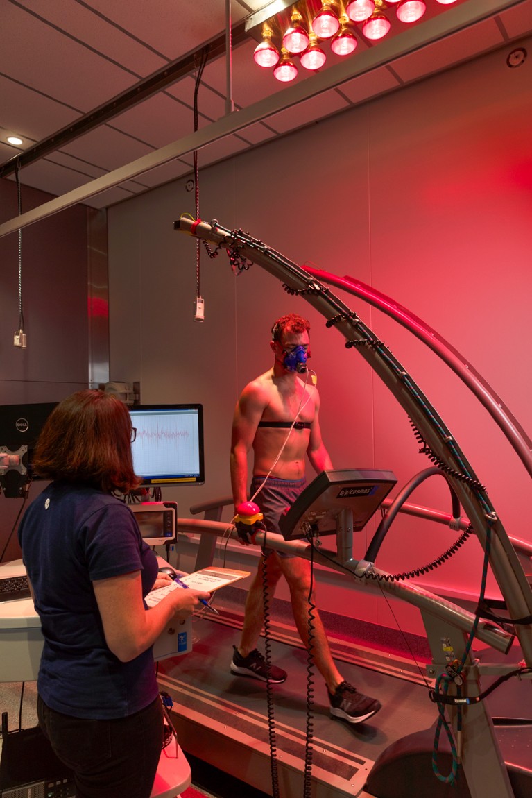 A researcher observes a man walking on a treadmill in climate chamber flooded with red light