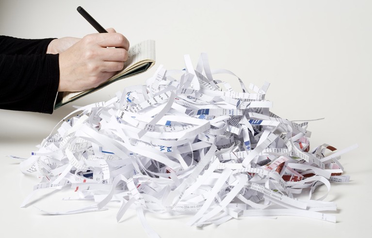 Close up view of hands writing on a notepad beside a pile of shredded paper documents.