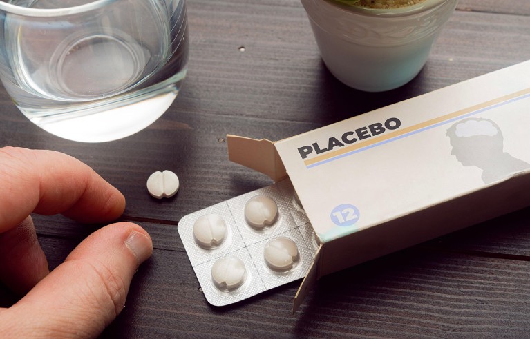 Close up view of a hand reaching to take a single white pill from beside a small box of pills labelled Placebo.