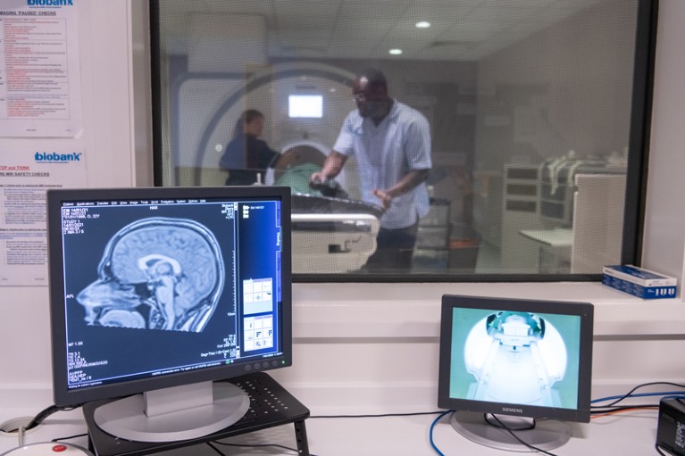 Technicians prepare a participant in an MRI scanner behind an MRI brain scan on a computer screen