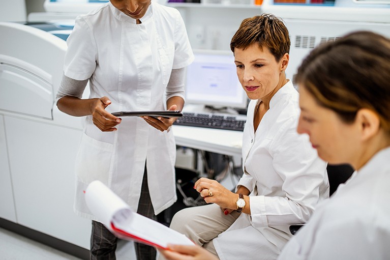 Three female scientists in white coats look at research papers in a laboratory.