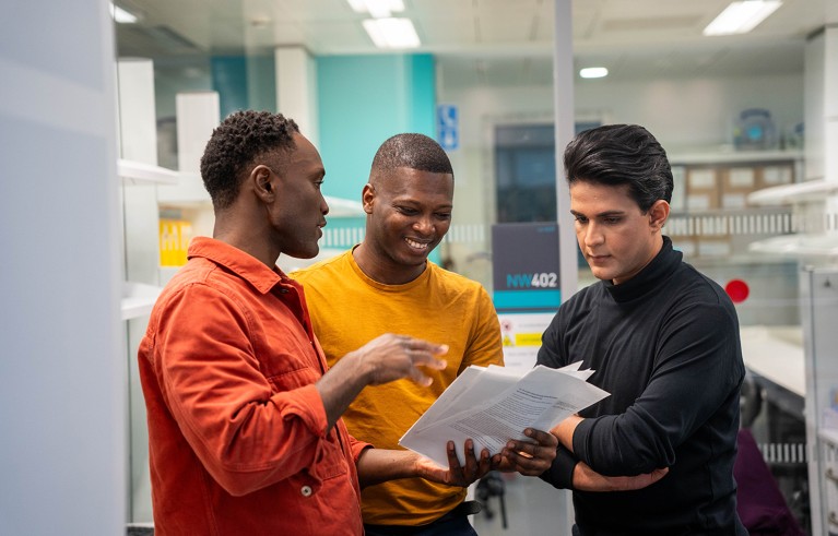 Three men (left to right) with an orange, yellow and black shirt talk over a paper in a research institute.