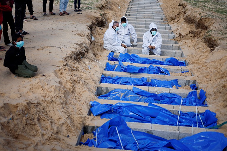 People in white coveralls sit by a mass grave as bodies are buried in blue bodybags.