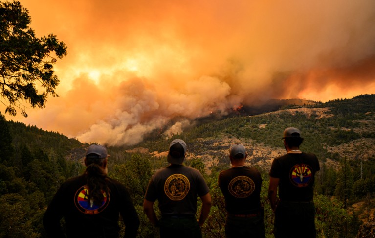 Vista de cuatro bomberos observando las llamas y el humo moverse hacia un valle en la distancia