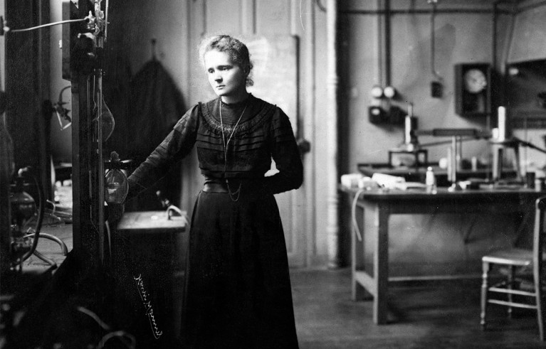 Black and white photo of Marie Curie standing in her laboratory surrounded by scientific instruments on tables