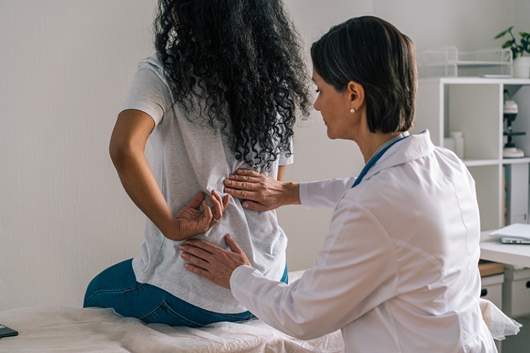 A female patient sits on a table while a female Physician examines her spine