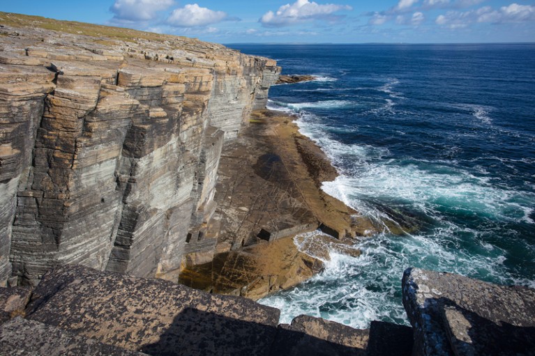 A rocky cliff edge on Orkney made of laminated Old Red Sandtone rock with waves crashing at the bottom of the cliff