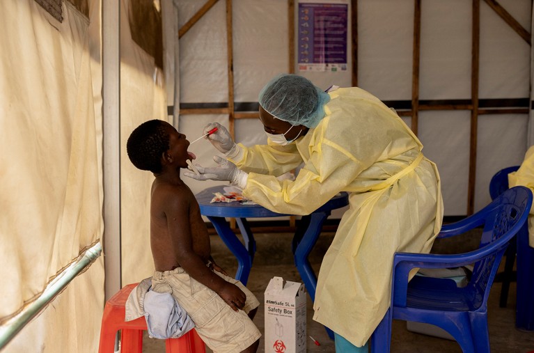 A laboratory nurse in protective clothing takes a mouth swab from a child.