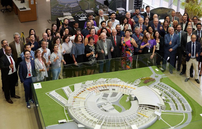 A crowd of people pose next to a model of a scientific facility.