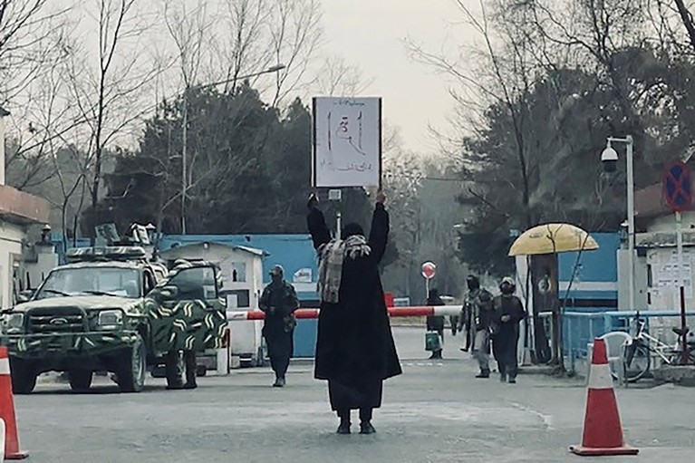 A lone student holds a protest sign against the ban on women's higher education outside the Kabul University as members of Taliban stand guard.