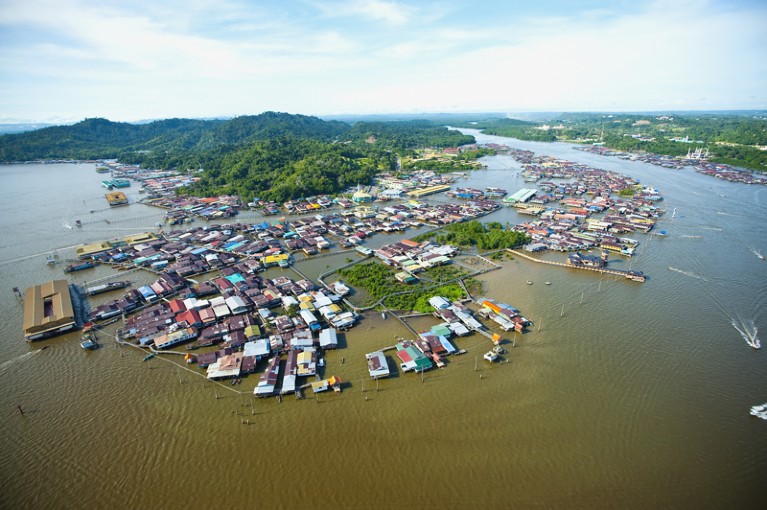 Aerial view of floating structures that make up Bandar Seri Begawan Floating Village, clustered in the water around forested hills in Brunei