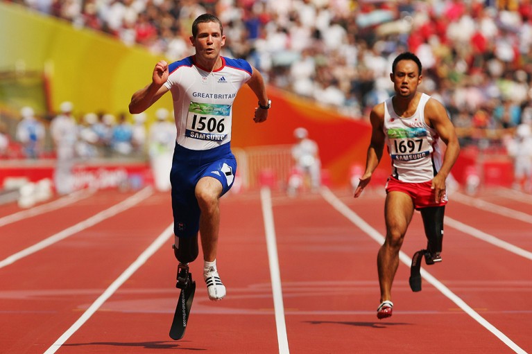 Two men, each with a prosthetic leg, sprinting on a track in a stadium.