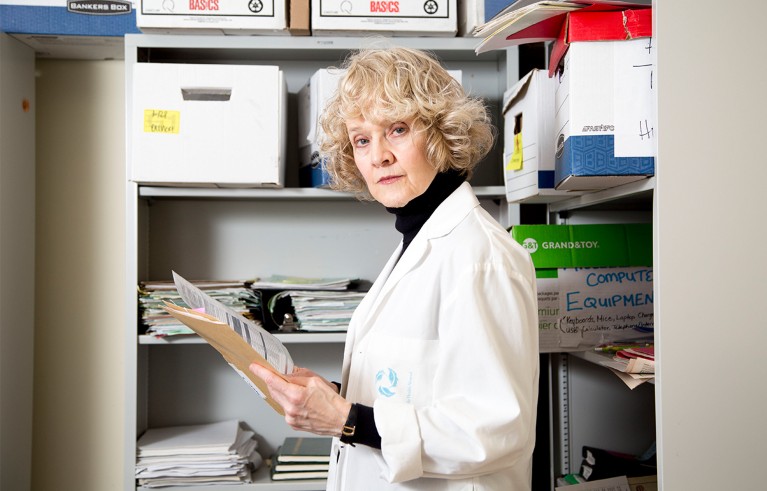 Woman in white coat standing in her office in front of a bookshelf holding some papers.