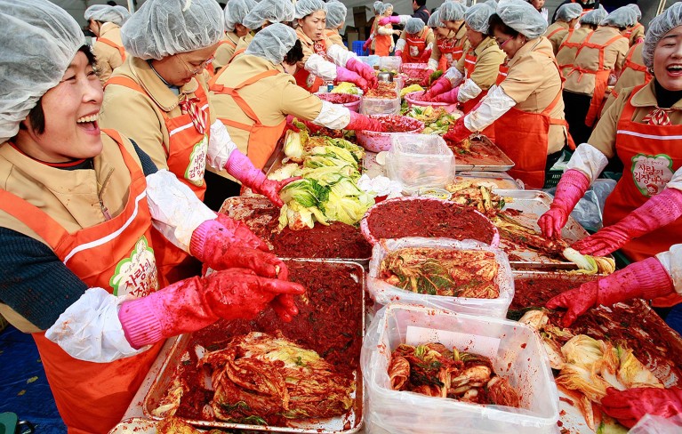 Many women pack around a table to make kimchi at a public event.