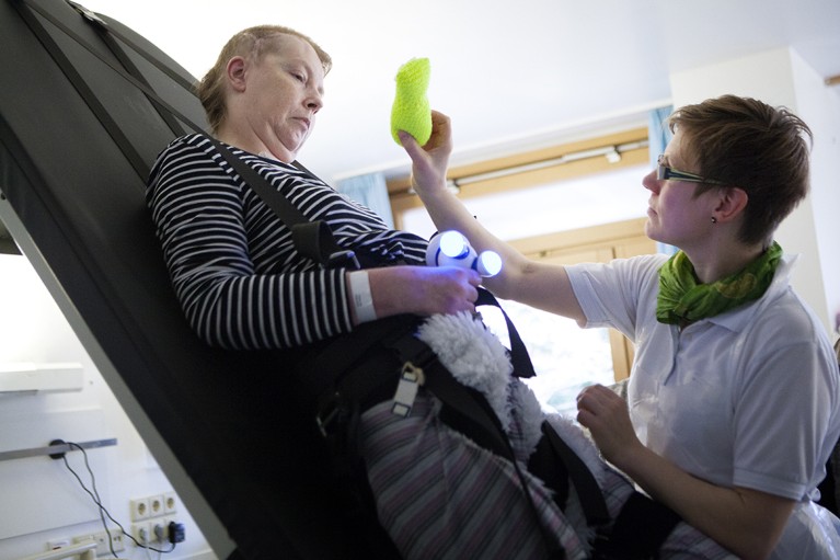 A woman strapped into a rehabilitation Erigo support system follows a bright green plushie with their eyes and grasps a light up plushie with their hands while a physiotherapist assists.