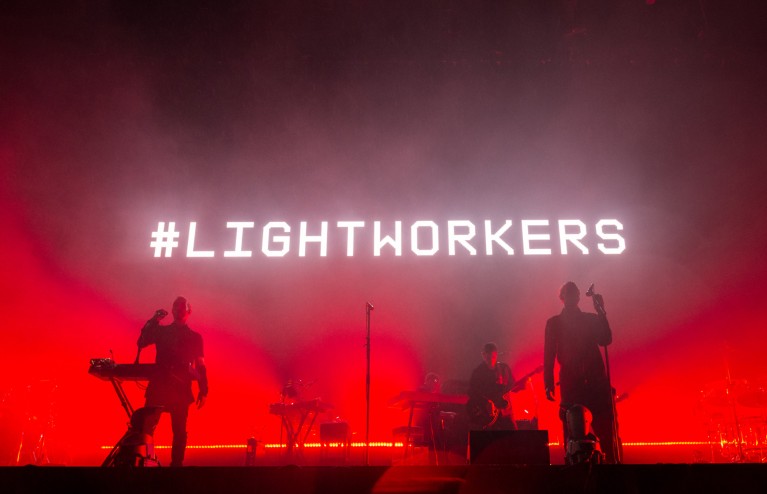 Close-up of band 'Massive Attack' performing on stage with a red screen behind them with white text reading #lightworkers