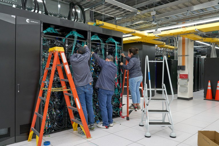 Technicians using stepladders examine the many wires and circuit boards behind panels of the Frontier Supercomputer