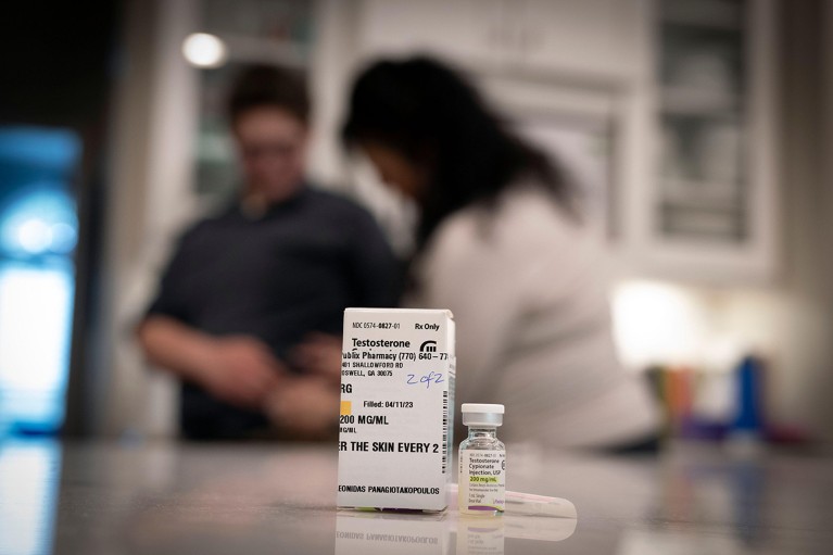 An injection of testosterone is given to a patient in the background, with the vial of testosterone in the foreground.