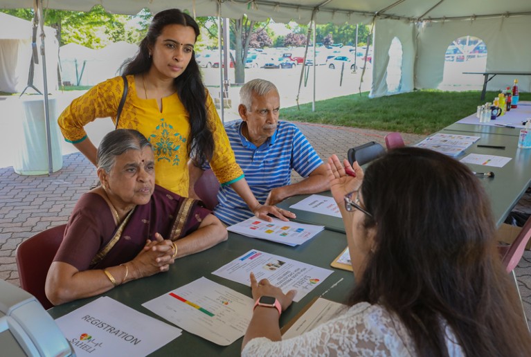 A researcher sat at a table in a marquee explains the SAHELI study to members of the South Asian community of Aurora, Illinois