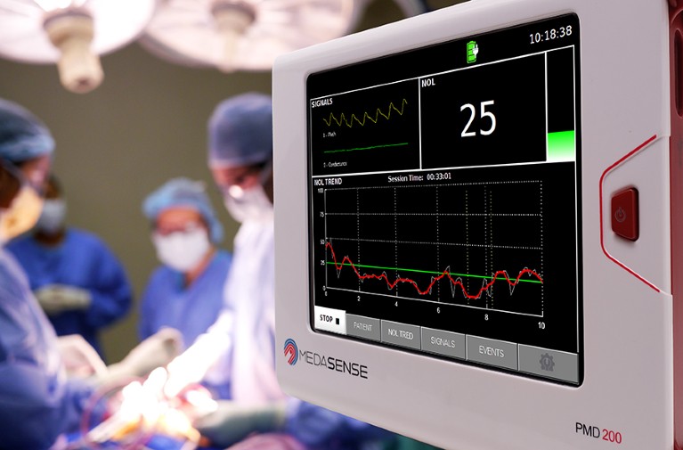 A digital screen with a steady green line and a wavering red line are showing in foreground, with a medical operation going ahead in the background out of focus