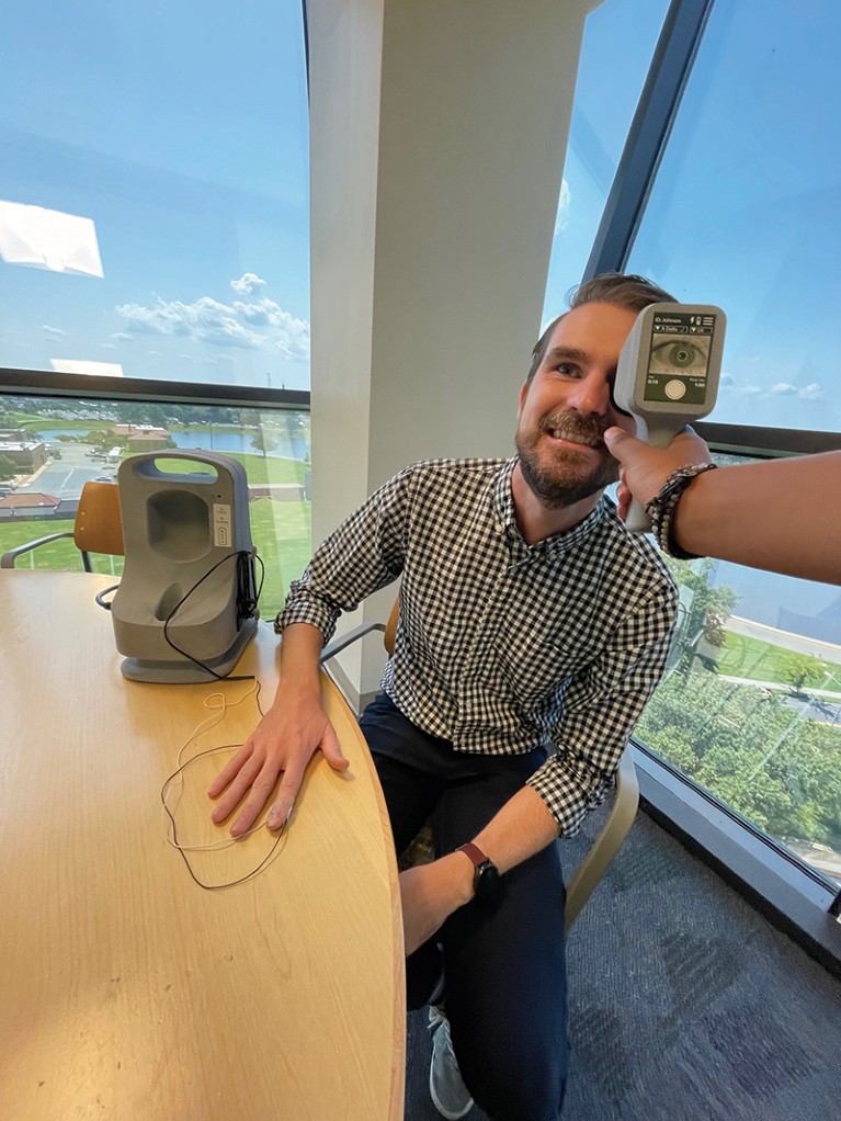 A closeup of a man’s eye is seen through the screen of a small handheld device. The man wears a pulse reader and sits in a high rise office.