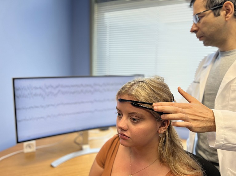 A woman wears an EEG device on her head; behind her results are streamed onto a screen and a researcher examines them