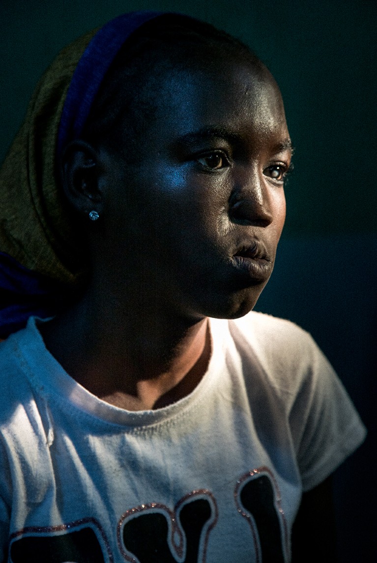 A Nigerian teenage woman in a colourful headscarf and tshirt with facial scarring from noma stares off camera