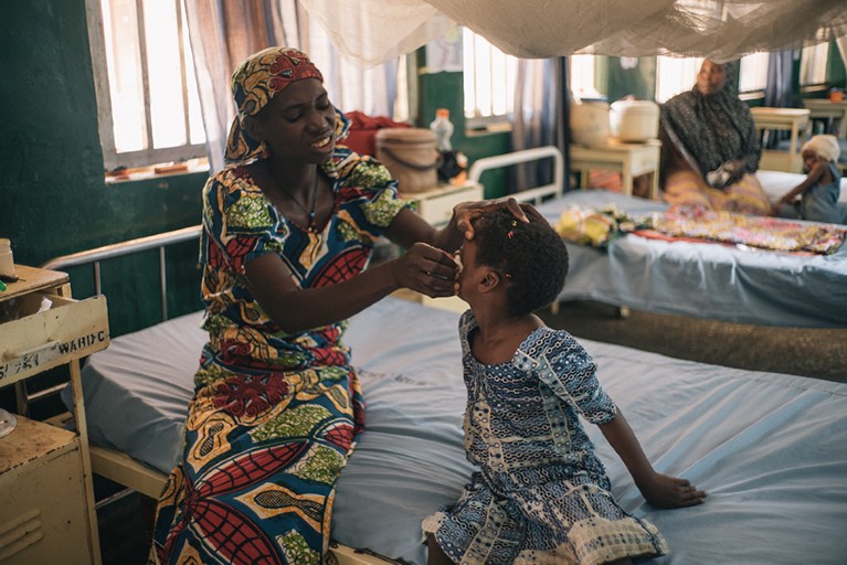 A Nigerian woman squints as she cleans the noma derived wound in her young daughter’s cheek while seated on a hospital bed in a ward. They are both wearing colourful Ankara dresses.