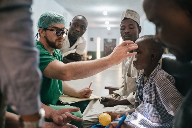 A doctor in scrubs reaches out to examine a young Nigerian boy in a check shirt with nasal disfigurement accompanied by his father (rear). Other medical staff are in the background and foreground.