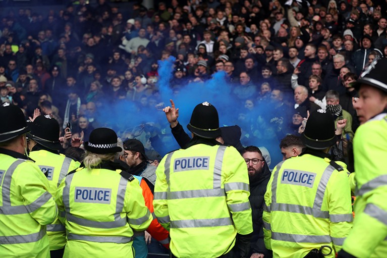 Police in high-vis jackets stand with their backs to the camera trying to control a crowd at a football match