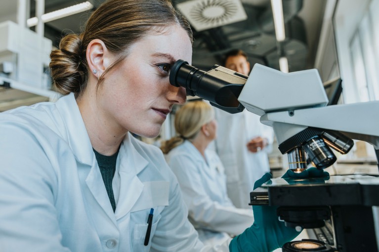 A female scientist in a white lab coat looks through a microscope in a laboratory with other scietists