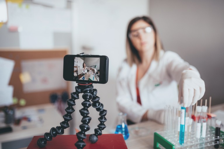 A female scientist in a lab coat using a phone on a tripod to record herself carrying out an experiment with test tubes