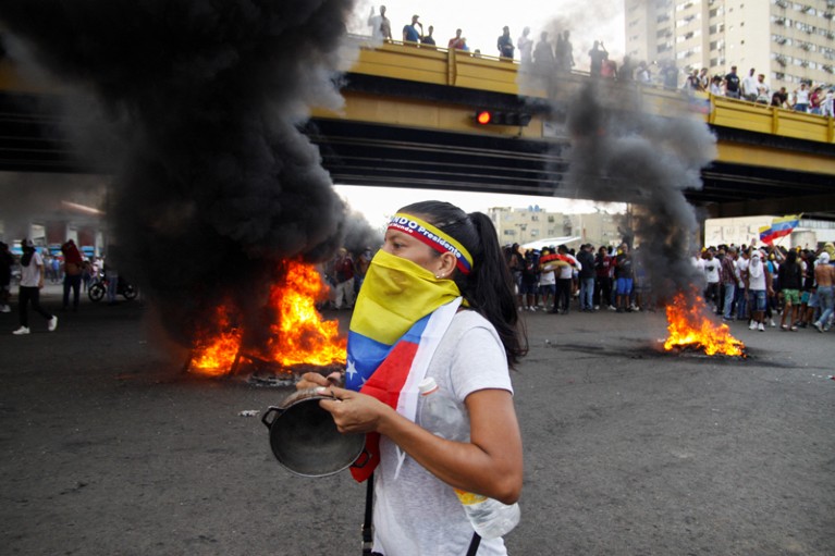 Las llamas arden detrás de una mujer que lleva una bandera venezolana en la cara y golpea una olla durante una protesta en Puerto La Cruz.