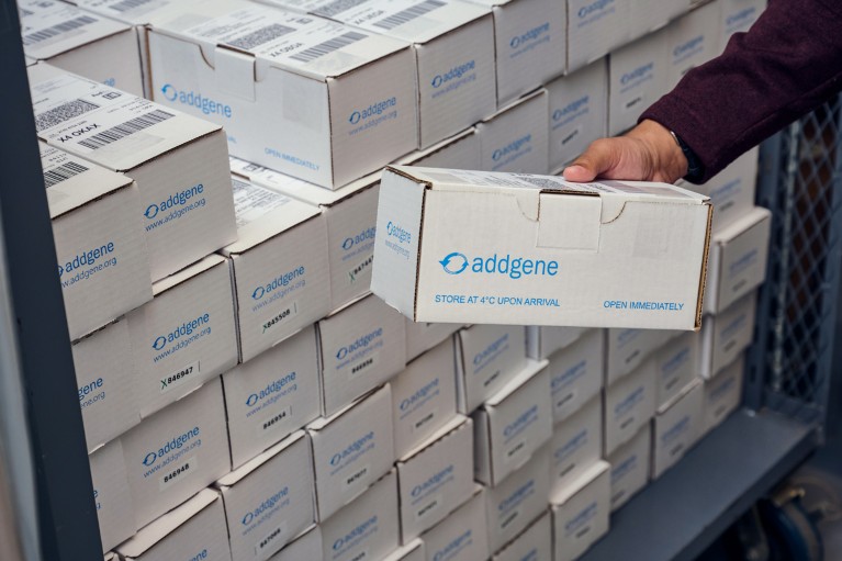 A hand picking up a box from a pile of white rectangular boxes with the blue Addgene logo printed on them