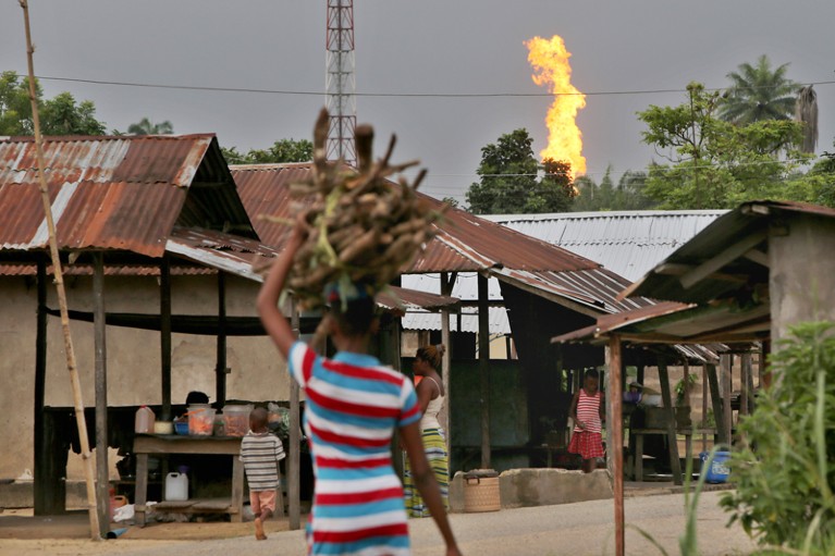 A woman carries a pile of firewood on her head through a street in Nigeria while a gas flare burns in the distance