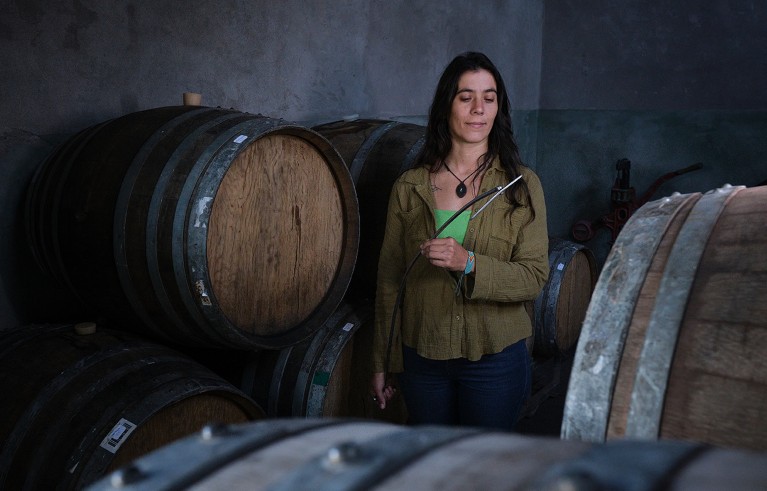 Mulher de longos cabelos escuros chamada Micaela Kuri retratada com a bengala realizando Battonage entre barris de vinho.