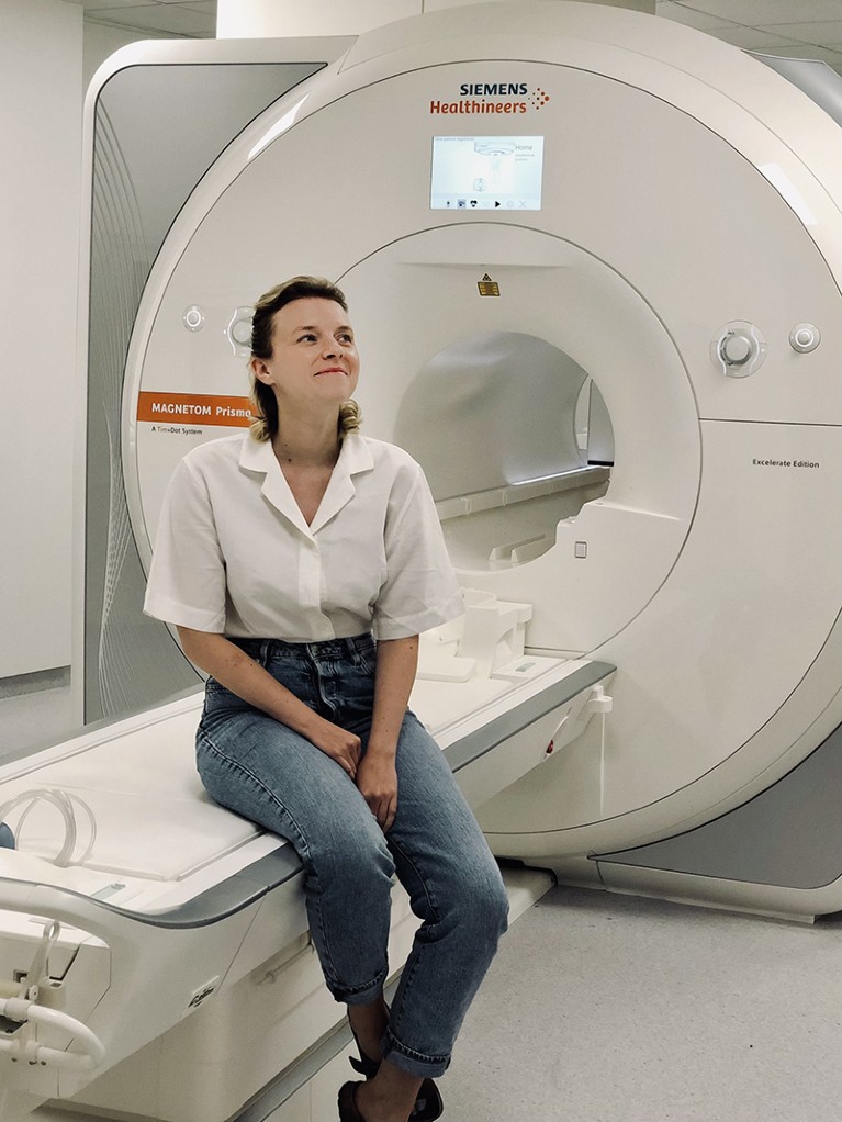 A photograph of Carina Heller sitting in front of an MRI machine