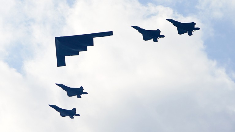 A B-2 Stealth Bomber flies alongside four F-35 fighter jets against a cloudy blue sky.