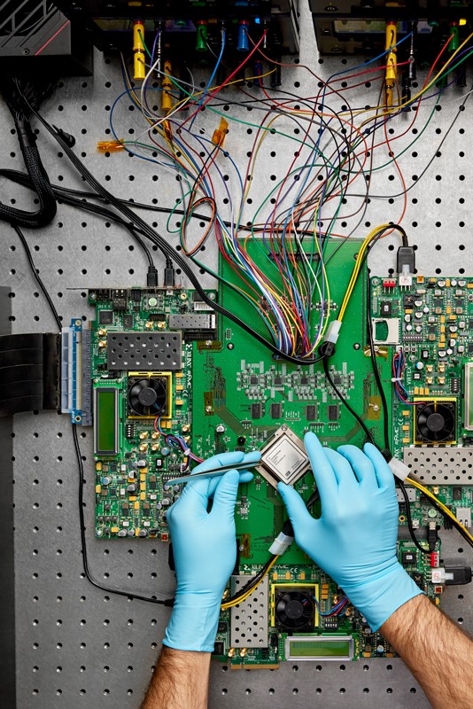 A top-down view of an analogue AI chip on a board with blue gloved hands holding tweezers