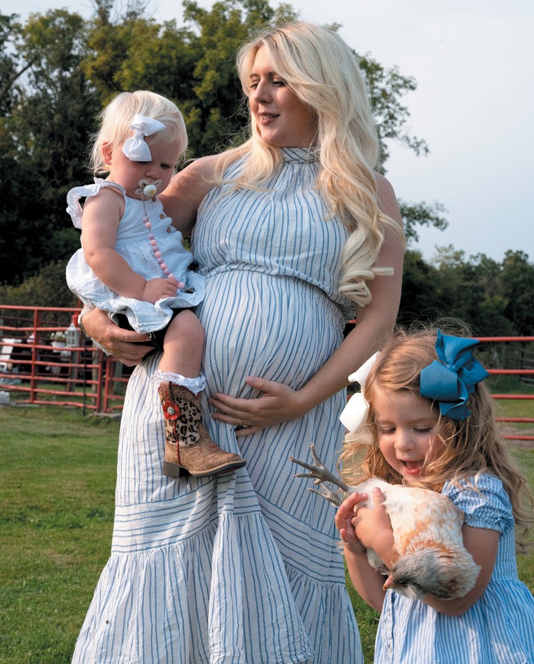 A woman holding a baby on her hip and a toddler standing below her, holding a chicken