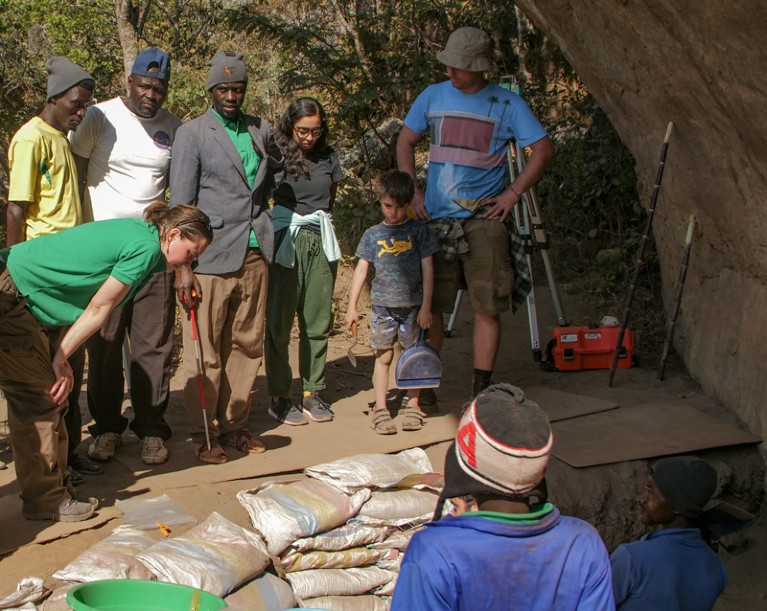 A group of visitors and field crew including Jessica Thompson look at some findings in an excavation pit while Jessica's son, aged 6, holding the trowel, looks on